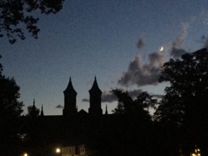 A dark silhouette of Antioch College's Main Hall against a blue sky with clouds and the moon visible in the background.
