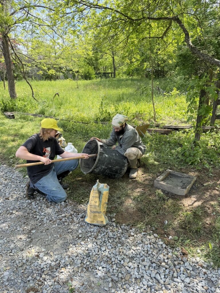 Two people crouch on a gravel driveway, one holding a plastic trash bucket, the other reaching into it with a shovel.